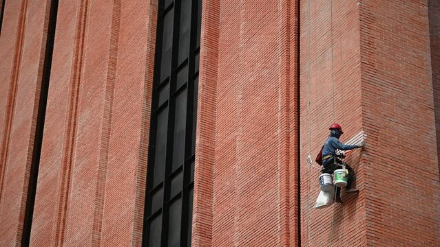 A builder is working at a height, holding on to a rope and safety net. The climber is painting the insulated facade of the building with a roller.