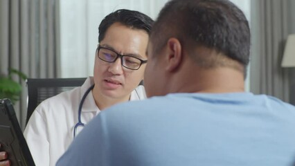 Close Up Of Asian Male Doctor With A Fat Male Patient In Clinic, Sitting At Desk, Discussing About The Results Of Medical Check Up On A Tablet, Giving Bad News, Encouraging A Patient
