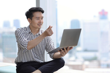Young asian businessman sitting and using computer while showing heart symbol in business office building and city background