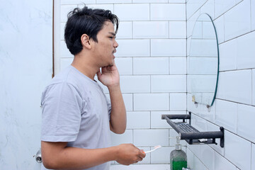 Side view of Asian man holding cheek suffer from toothache while looking at mirror in the bathroom