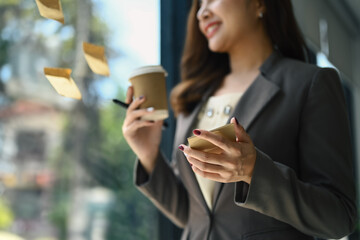 Businesswoman executive manager holding paper cup of coffee and reading sticky notes on glass window