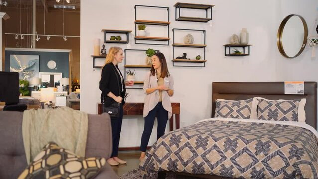 A Young female sales associate person leads a shopping customer to a bedroom display to demonstrate the Comfortable Mattress before leaving inside a retail furniture store