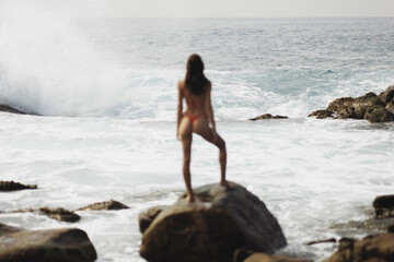 Epic extreme scene of big ocean waves crashing on rocky coastline in front of bikini beach model. High quality photo