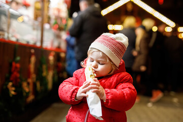 Little baby girl, cute child eating bananas covered with chocolate, marshmellows and colorful sprinkles near sweet stand with gingerbread and nuts. Happy toddler on Christmas market in Germany.