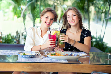 Two happy teenage young girls met each other in the cafe drinking fruit cocktails. Summer warm day on vacations.