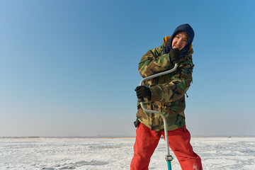 A warmly dressed Asian man drills a hole in the ice on winter fi