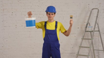 Medium shot of a young construction worker standing in the room under renovation, showing a bucket full of paint for walls and a brush.