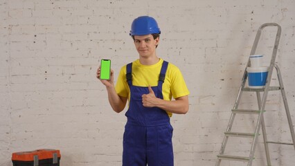 Medium shot of a young construction worker standing in the room, holding a smartphone with advertising area, workspace mock up and giving a thumbs-up.