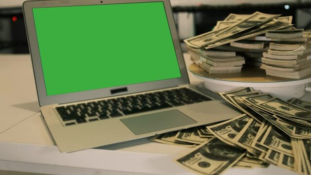 Open laptop green screen with row of scattered 100 dollar bills and packs of hundred banknotes folded on rotating surface. Online winning and cash flow.