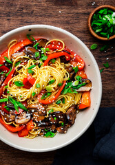 Stir fry noodles with vegetables: paprika, champignons, chives and sesame seeds in ceramic bowl. Wooden table background, top view
