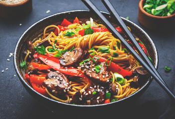 Stir fry noodles with vegetables: red paprika, champignons, green onion and sesame seeds in ceramic bowl. Black table background, top view