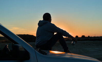 A man sits on the hood of a car in front of the sun at sunset. A man sits conceived with a thermo...