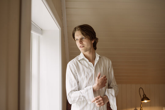 Happy young man standing near window and buttoning shirt sleeves