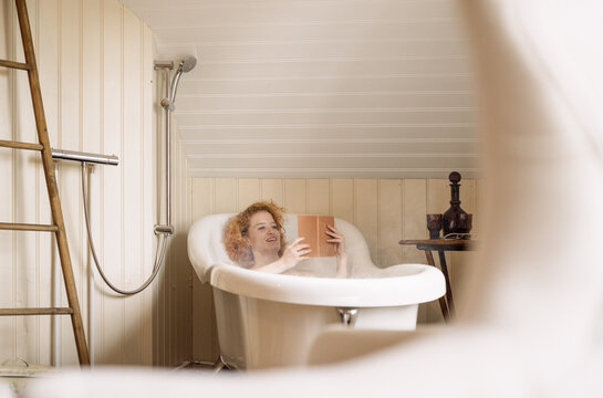 Young woman lying in bathtub and reading book in daylight