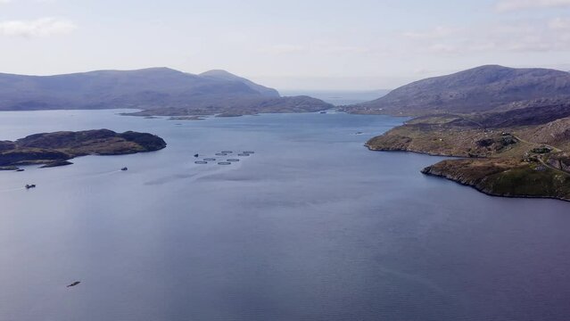 Wide drone shot of the bay around Tarbert, a village on the Isle of Harris, part of the Outer Hebrides of Scotland. A fish farming facility is visible as well as many mountains and cliffs.