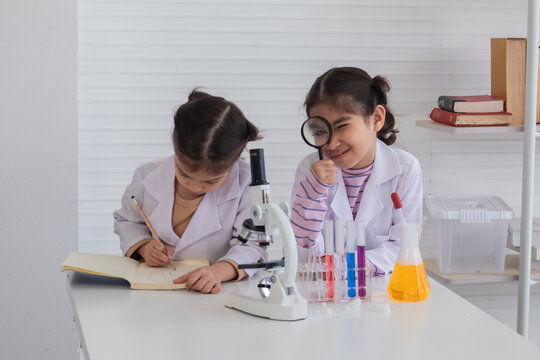 Cheerful Arabian student girls wearing safety eyeglasses using magnifier glasses and microscope in lab coat uniform, sibling girls play roles together in science class at school, future kid scientist
