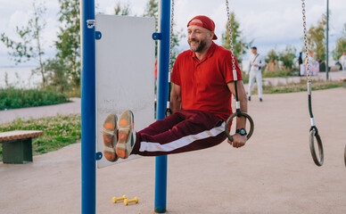 Cheerful mid aged caucasian man in sportswear training at sport court lifts legs leans on gymnastic rings at park. Strong bearded European guy at exercise outside. Healthy people, sport, CrossFit.