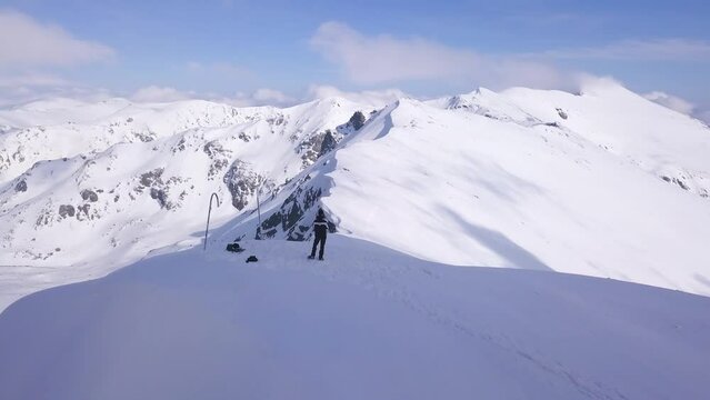 Man stands alone on peak of Parang Mountain with snowy landscape background. Aerial view from drone flying along ridge overhead.