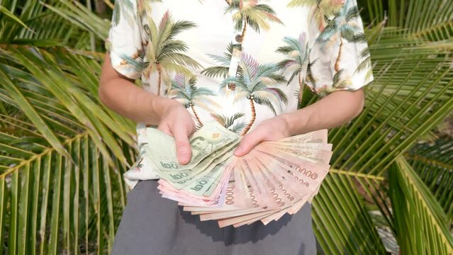 Woman in a tropical shirt counting Thai Baht on a sunny day with palm fronds in the background. Close up.