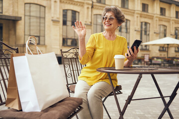 Senior woman with smartphone sitting at cafe terrace while drinking coffee and waving hi to friends