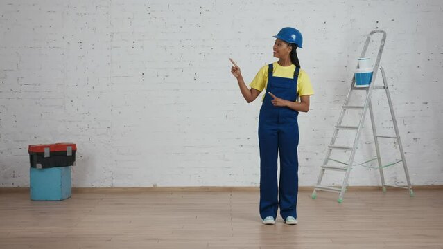 Full length video of a dark-skinned young female construction worker standing in the room under renovation pointing to the side of the frame. Place for your advertisement.