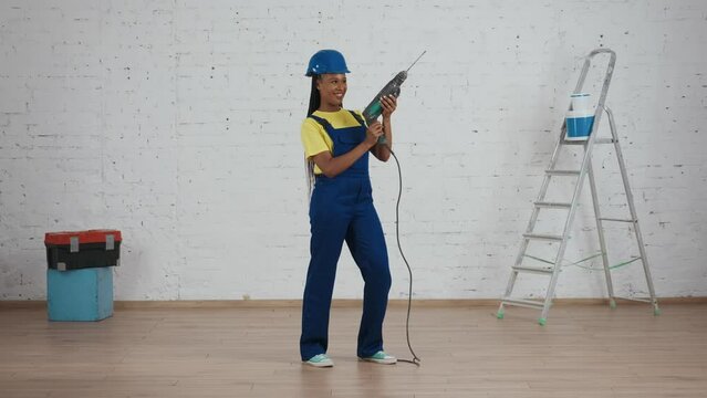 Full length video of a dark-skinned young female worker standing in the room under renovation, looking around, raising the drill, perforator and holding it like a gun.