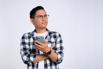 Pensive young Asian man in casual shirt holding smartphone, looking aside at copy space, thinking about offer isolated on white background