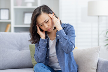 Sick woman alone at home, Asian depressed woman having severe headache, sitting on sofa in living...