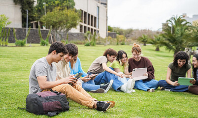 Happy students studying with digital tablets sitting on grass at campus park with college building...