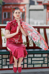 A young Asian girl walks in national clothes with a red umbrella in the park. Chinese woman in a red traditional dress, close-up portrait