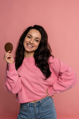 Cheerful pretty lady with an oat biscuit in one hand posing for the camera on the pink background. Self-indulgence and unhealthy food concept