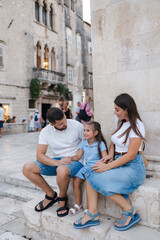 Young family of dad mom and daughter sits on stone in old town. Beautiful people. Denim style