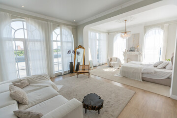 cozy bright apartment with huge panoramic windows with sunlight. bedroom in white and beige tones with fireplace and soft armchair. stylish living room with log coffee table and large white sofa