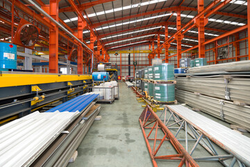 A gargantuan warehouse brims with a labyrinth of gleaming steel beams crisscrossing overhead, creating a metallic canopy. There are steel sheet roll and metal sheets on the floor.