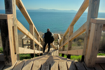 A man with a backpack goes down a wooden staircase going sharply down to the sea on a clear sunny day