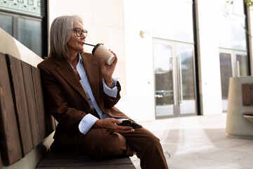 charming successful senior woman with gray hair and glasses dressed in a brown designer suit drinking coffee on the street