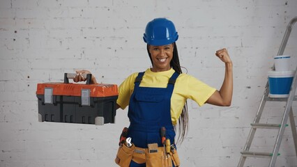 Medium shot of a dark-skinned young female construction worker standing in the room, wearing a tool belt, raising a tool box and showing her muscles.
