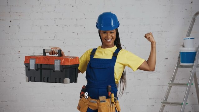 Medium shot of a dark-skinned young female construction worker standing in the room, wearing a tool belt, raising a tool box and showing her muscles.