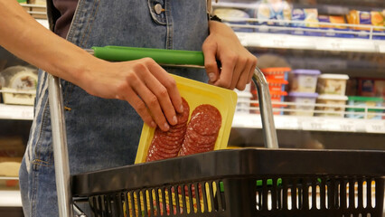Close-up of a young woman's hands putting a package of sliced salami into a shopping cart