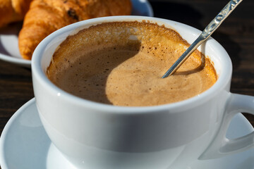 Thick foam of hot coffee cappuccino with spoon in white cup and croissant on wooden table, closeup. Milk foam cappuccino, latte