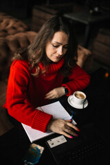 woman in a red sweater works in a coworking cafe with a laptop and a notebook, freelance work in winter, work on the Internet