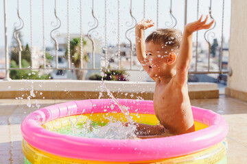 Toddler boy gleefully plays in an inflatable round swimming pool on the balcony during a hot summer day