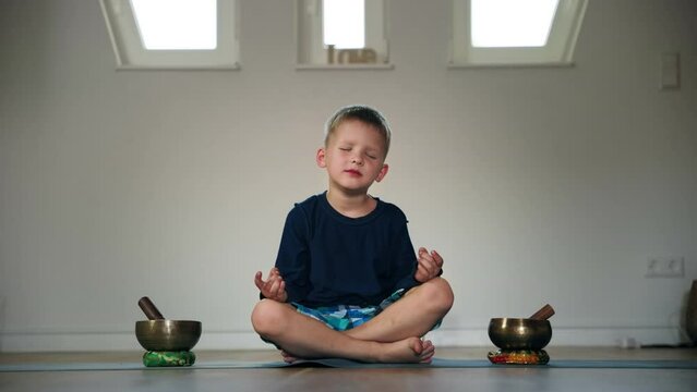 yoga for children, cute boy concentrated on meditation sits in lotus position on karimat near tibetan musical bowls in a bright room