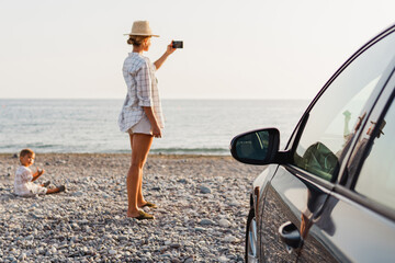 Woman traveler stands beside her car on a pebble beach and capturing the scenic beauty with her smartphone