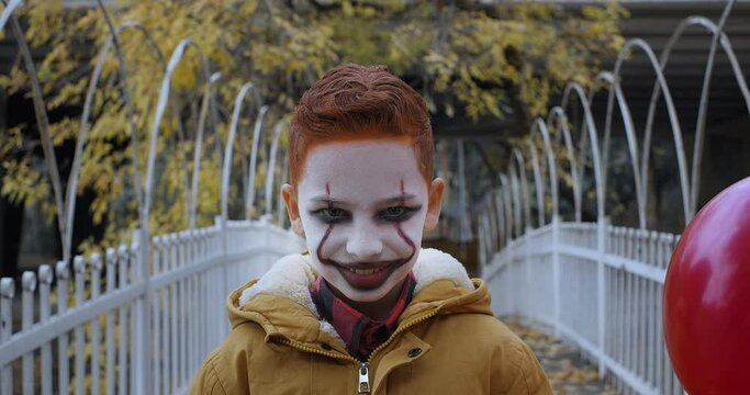 The boy in scary clown make-up looking into the camera and smiling ominously. Halloween costume party, horror movie concept