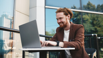  A handsome red-haired businessman is sitting on the summer terrace of the business center of the city. Happy man in a business suit is smiling.