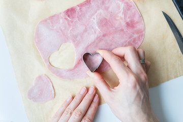 The cook cuts heart-shaped sausage-ham cut into heart shape on parchment, top view, close-up....