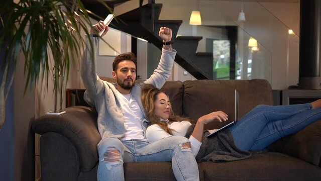 A man celebrating a goal on the sofa at home with his girlfriend who is with the computer, lifestyle of a couple on a romantic evening