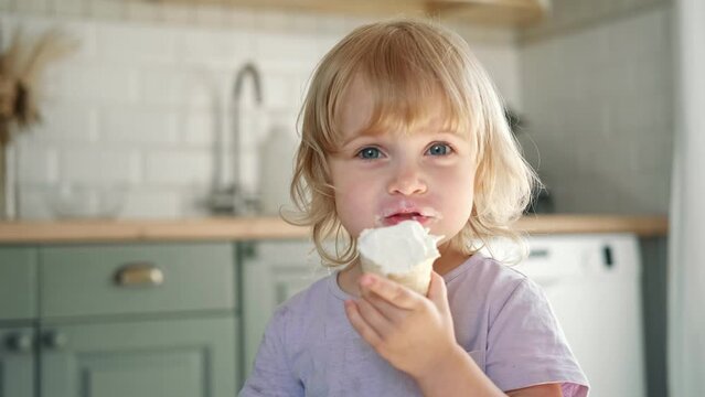 Baby girl enjoying ice cream. Pretty little toddler eating an ice-cream indoors, at home. Dining room background. Small child eats plombir and cream messy on her mouth. Cute kid with tasty sweet food.