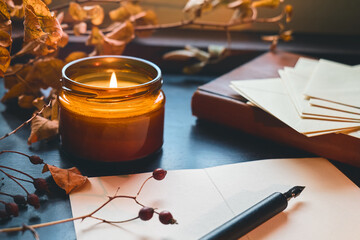 Cozy autumn still life fall leaves, sheets of paper, fountain pen and burning candle on the windowsill. Autumn home decor. Cozy fall mood.